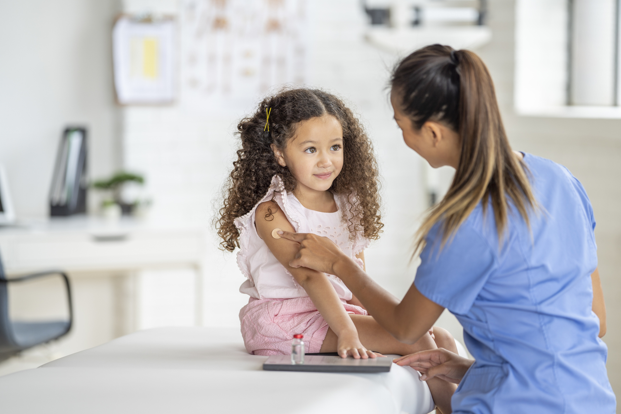 Healthcare professional placing a bandage on a young child’s upper arm after an immunization while the child sits on an examination table in a medical office.