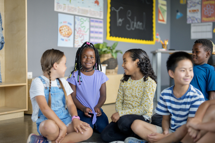 Group of elementary-age children seated together on a classroom floor during a collaborative learning activity, with educational materials and a chalkboard visible in the background.