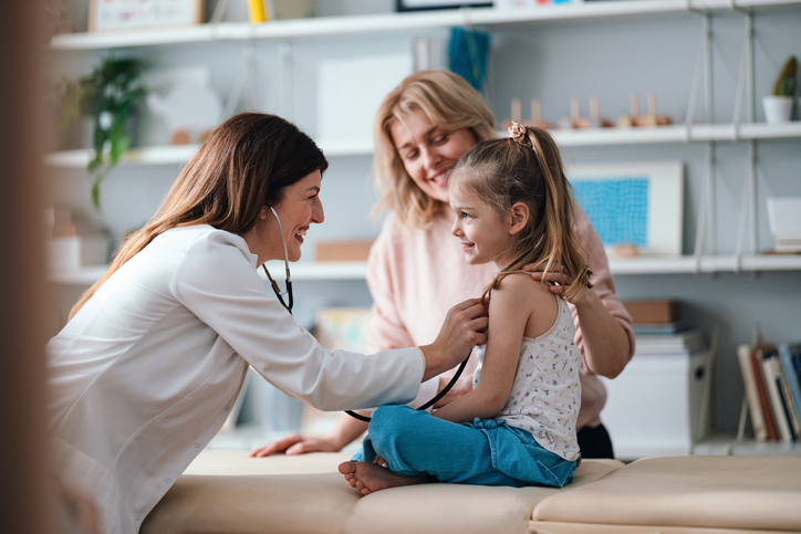 Healthcare professional using a stethoscope to examine a young child during a routine checkup while another adult sits nearby in a clinical setting.