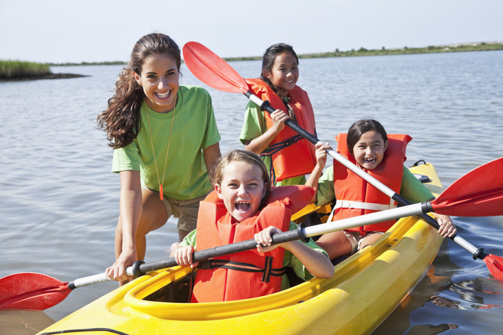Teenage camp counselor is smiling while she helps three children on a kayak enjoying summer camp.