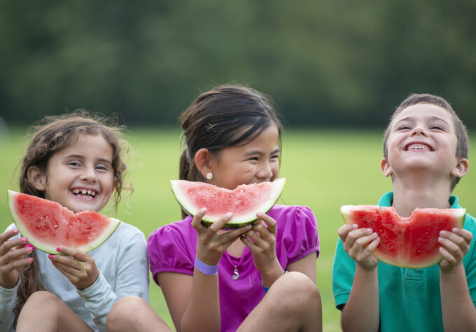 Three school aged children hold watermelon slices up in front of them as they smile for a portrait.