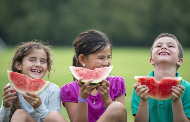 Three school aged children hold watermelon slices up in front of them as they smile for a portrait.