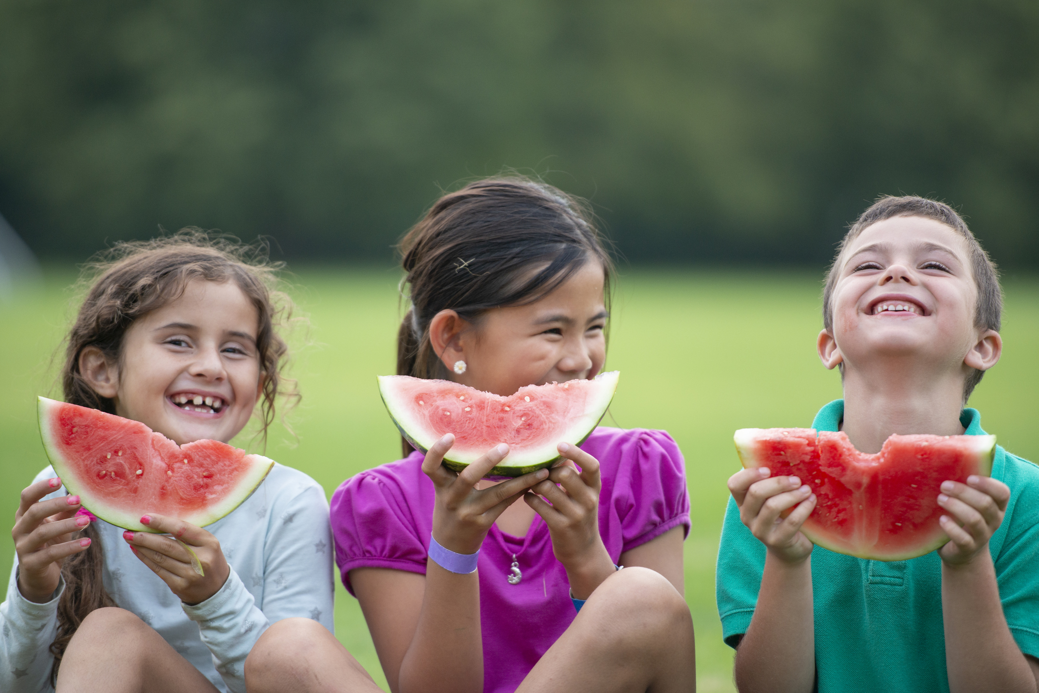 Three school aged children hold watermelon slices up in front of them as they smile for a portrait.