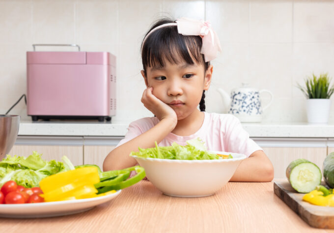 Child sits at a table in front of fresh vegetables with a thinking expression wondering if she should eat it.