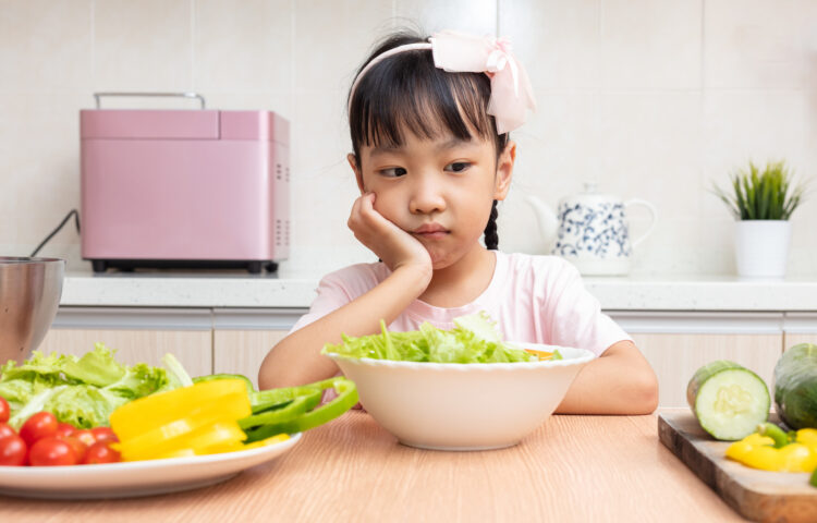 Child sits at a table in front of fresh vegetables with a thinking expression wondering if she should eat it.