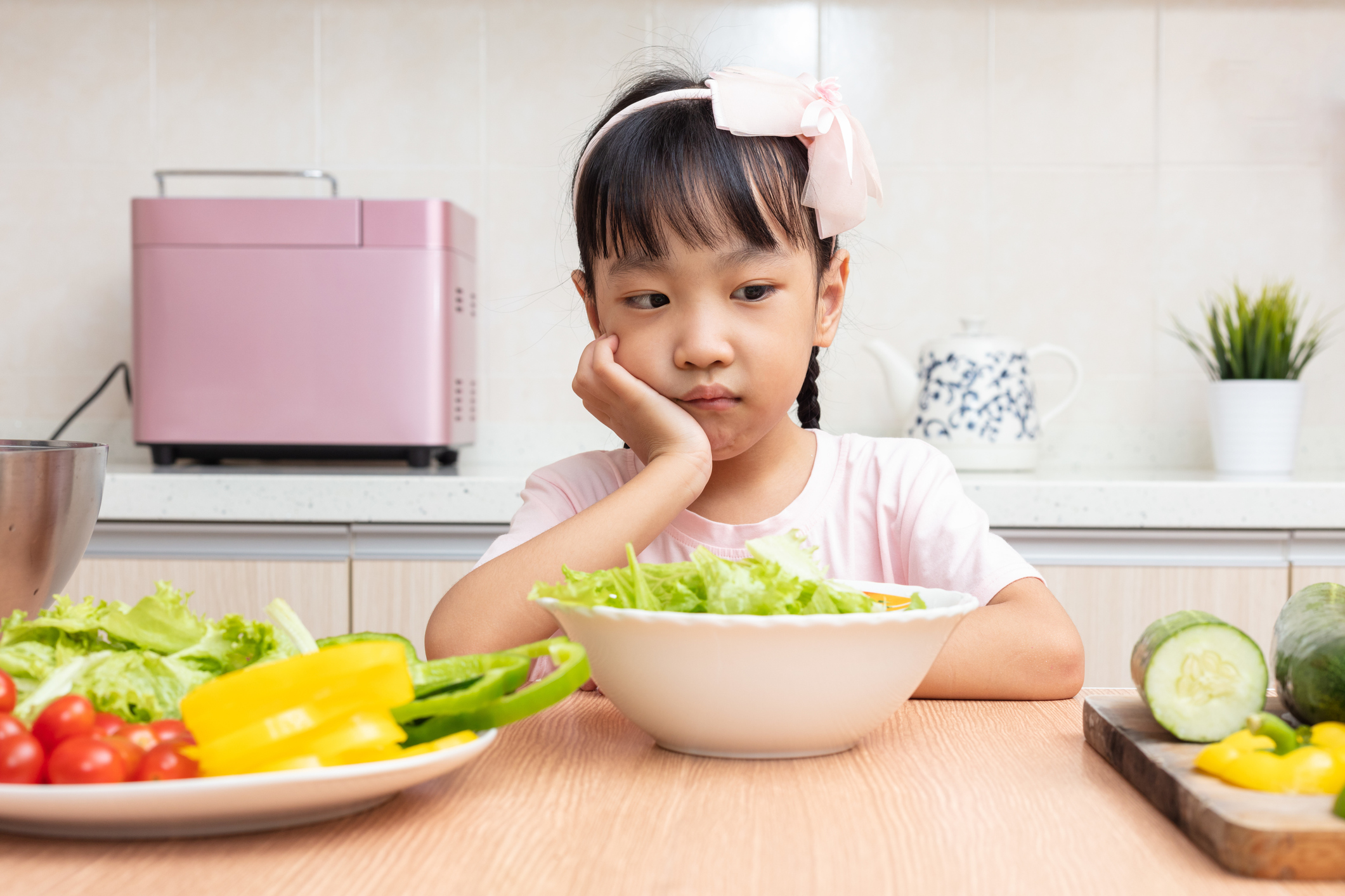 Child sits at a table in front of fresh vegetables with a thinking expression wondering if she should eat it.