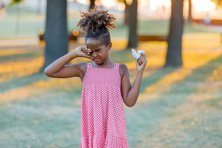 Child outdoors rubbing one eye and holding a tissue, showing common symptoms associated with seasonal allergies.