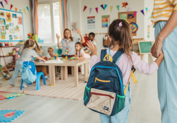 Young child wearing a backpack enters an early childhood classroom while holding an adult's hand as other children participate in learning activities.