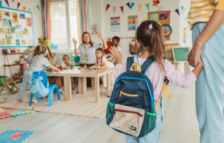 Young child wearing a backpack enters an early childhood classroom while holding an adult's hand as other children participate in learning activities.