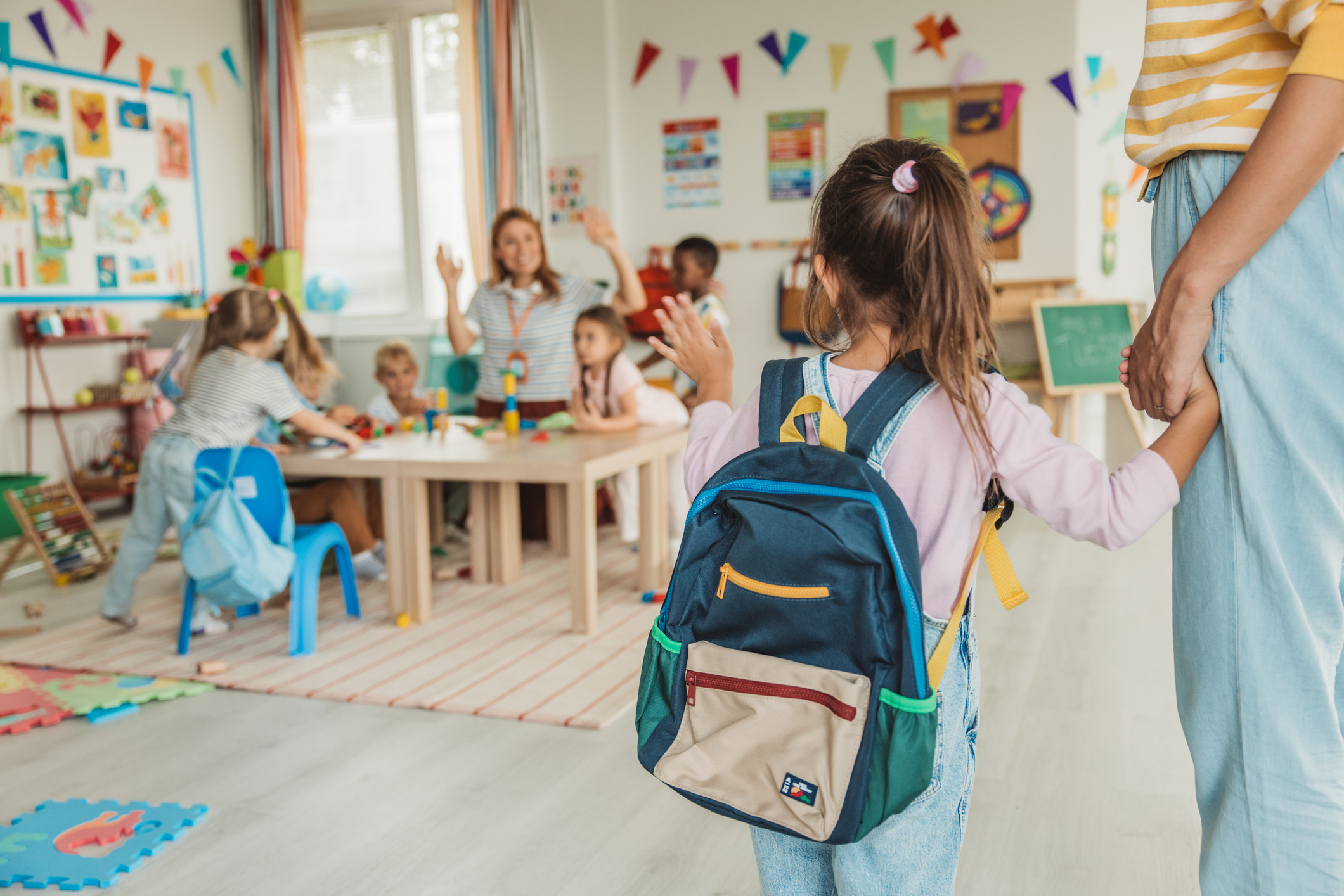 Young child wearing a backpack enters an early childhood classroom while holding an adult's hand as other children participate in learning activities.