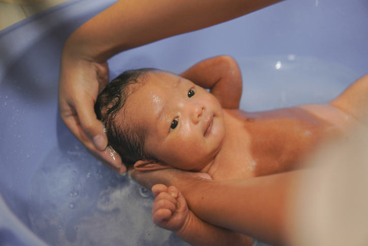 Calm newborn baby is being washed in a bathtub by parent.