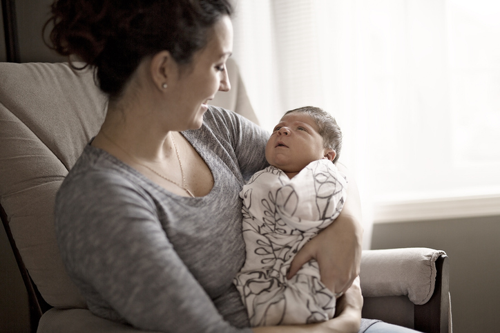 A smiling mother sitting in a chair holding a swaddled newborn baby who is gazing up at her.