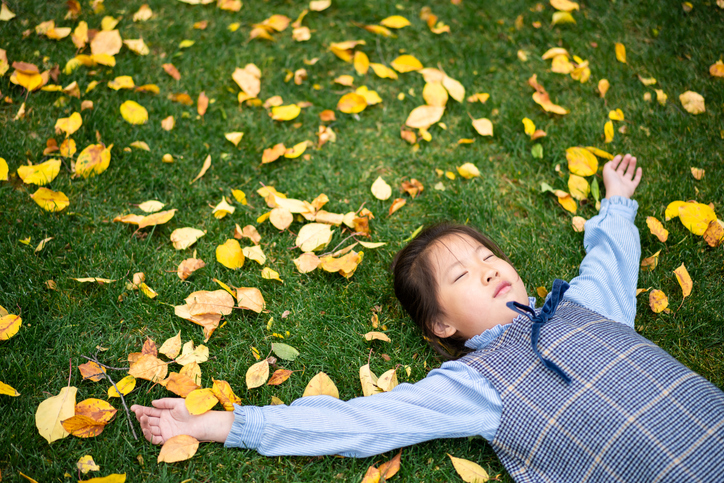 Young child lying on grass covered with autumn leaves, representing the seasonal transition that accompanies the fall time change.