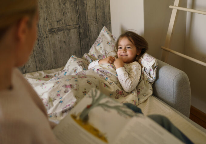 Child lying in bed while an adult reads a bedtime story as part of a consistent nighttime routine.