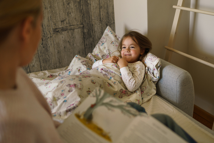 Child lying in bed while an adult reads a bedtime story as part of a consistent nighttime routine.