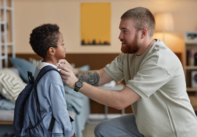 Child wearing a backpack stands with an adult in a bedroom while settling into a new home.
