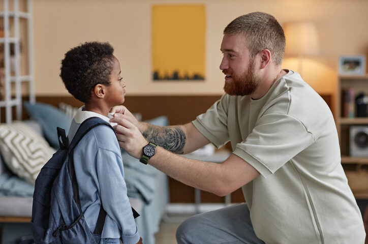 Child wearing a backpack stands with an adult in a bedroom while settling into a new home.