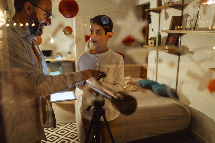 Adult and child use a telescope together in a bedroom decorated with personal belongings.